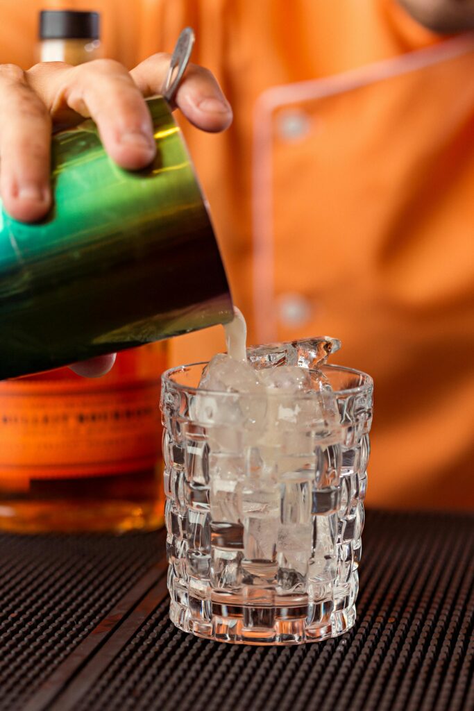 pexels-photo-14161978-14161978 Close-up of a bartender pouring a cocktail into a glass with ice cubes at a bar counter.